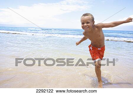 Stock Image - A young boy runs and plays in the shallow water of the ocean; Hawaii united states of america. Fotosearch