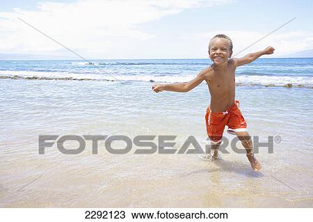 Stock Image - A young boy runs and plays in the shallow water of the ocean; Hawaii united states of america. Fotosearch
