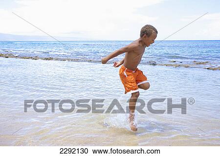 Stock Image - A young boy runs and plays in the shallow water of the ocean; Hawaii united states of america. Fotosearch