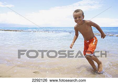 Stock Photograph - A young boy runs and plays in the shallow water of the ocean; Hawaii united states of america. Fotosearch