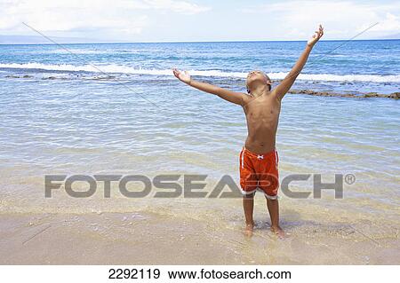 Stock Photo - A young boy standing in the shallow water of the ocean with arms raised and looking up; Hawaii united states of america. Fotosearch