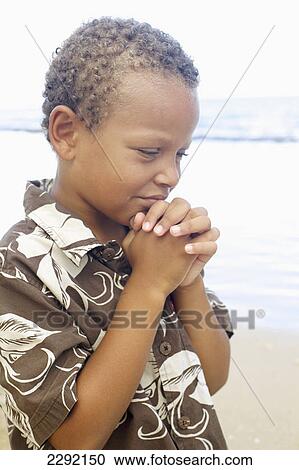 Stock Image - A young boy with hands folded in prayer by the ocean; Hawaii united states of america. Fotosearch