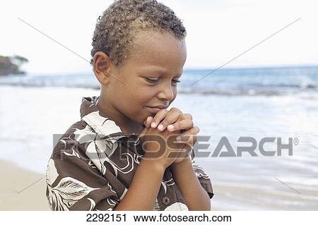 Stock Image - A young boy with hands folded in prayer by the ocean; Hawaii united states of america. Fotosearch