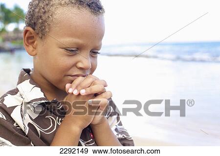 Stock Photo - A young boy with hands folded in prayer by the ocean; Hawaii united states of america. Fotosearch