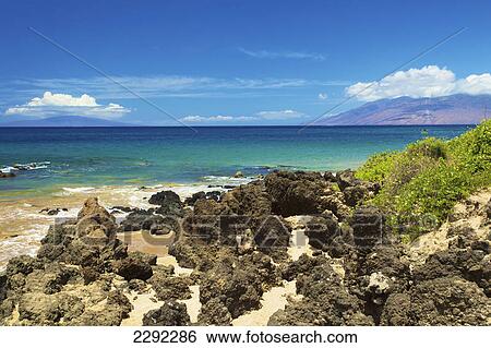 Stock Photograph - Rugged rocks sitting on the sand on the shore of an hawaiian island with mountains along the coastline; Hawaii united states of america. Fotosearch