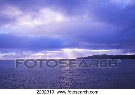 Stock Image - Sun rays reflected on the water at sunset with the coastline of an hawaiian island; Hawaii united states of america. Fotosearch