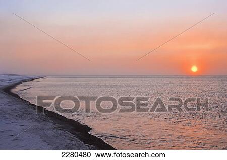 Stock Image - The sun shines through windblown snow above the beaufort sea coast just before it sets outside of barrow in winter; Alaska united states of america. Fotosearch