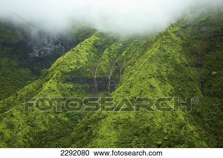 Stock Image - Waterfalls flowing through lush green hills under the low lying clouds; Hawaii united states of america. Fotosearch