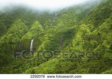 Stock Photograph - Waterfalls flowing through lush green hills under the low lying clouds; Hawaii united states of america. Fotosearch