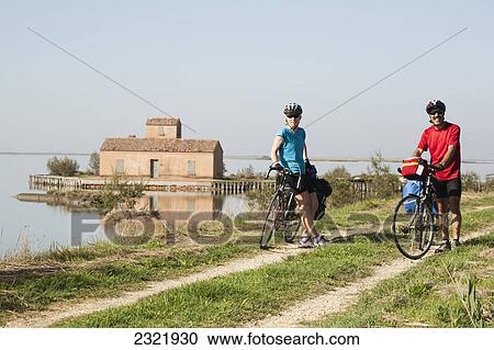 A Couple With Touring Bikes Along A Path And Body Of Water With A Boathouse In The Background; Comacchio Emilia-Romagna Italy View Large Photo Image Stock Image - A Couple With Touring Bikes Along A Path And Body Of Water With A Boathouse In The Background; Comacchio Emilia-Romagna Italy. Fotosearch