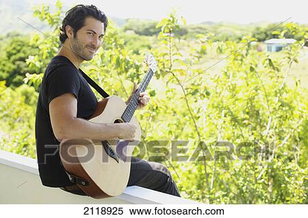 Stock Photography - A Man Playing His Guitar While Sitting On A Balcony Ledge With A View Of The Lush Vegetation; Wailua Kauai Hawaii United States Of America. Fotosearch