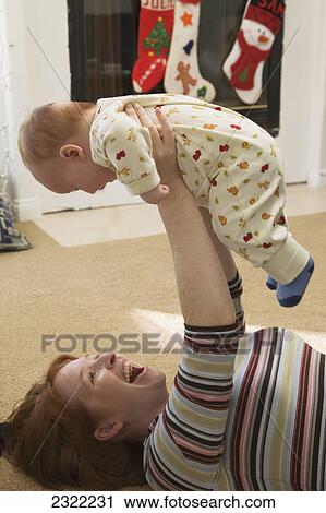 Stock Image - A mother laying on the floor and playing with her infant son with christmas stockings hung over the fireplace in the background; San diego california united states of america. Fotosearch