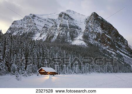 Stock Photo - A snow covered log cabin on a lakeshore with lights at dusk surrounded by snow covered evergreen trees and mountains; Lake louise alberta canada. Fotosearch