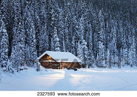 Stock Image - A snow covered log cabin on a snow covered lakeshore surrounded by evergreen trees at dusk; Lake louise alberta canada. Fotosearch