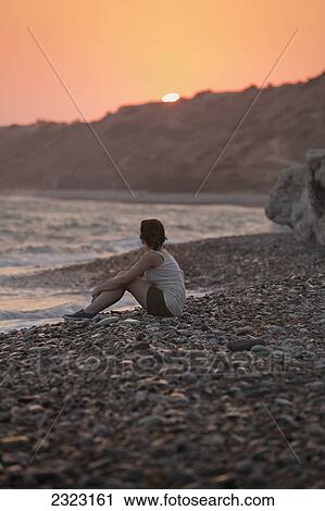 Stock Image - A woman sits on the rock beach looking out to the ocean at sunset; Aphrodite bay cyprus. Fotosearch