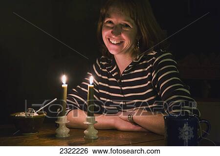 Stock Photograph - A woman sitting at a table in a dark room with a coffee cup and a bowl of ice cream being illuminated by two candles in front of her; Willimantic connecticut united states of america. Fotosearch