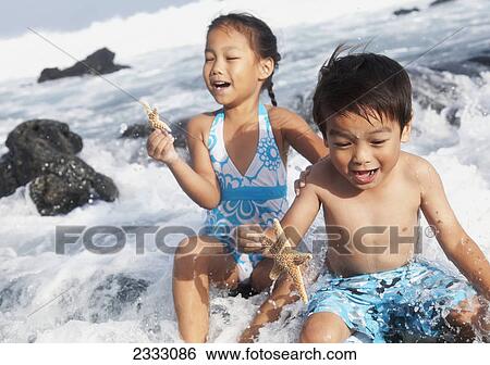 Stock Photograph - A young boy and girl hold starfish and sit in the crashing waves at the water's edge; Honolulu oahu hawaii united states of america. Fotosearch