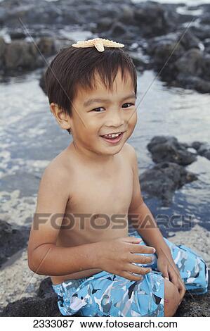 Stock Photo - A young boy sits at the water's edge with a starfish on his head; Honolulu oahu hawaii united states of america. Fotosearch