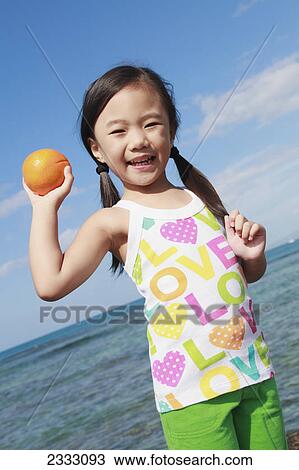 Stock Image - A young girl holds an orange and poses for the camera at the water's edge; Honolulu oahu hawaii united states of america. Fotosearch
