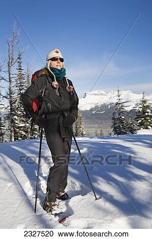 Stock Image - Female snowshoer in the mountains with toque and backpack and poles with snow covered mountains and blue sky; Lake louise alberta canada. Fotosearch