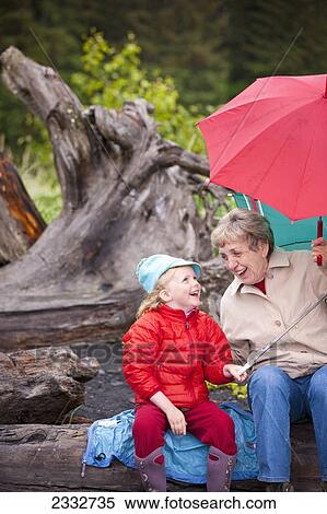 Grandmother and granddaughter playing with their umbrella while sitting on a log at the beach at Lowell Point on a rainy day, summer, Seward, Southcentral Alaska, USA View Large Photo Image Stock Photography - Grandmother and granddaughter playing with their umbrella while sitting on a log at the beach at Lowell Point on a rainy day, summer, Seward, Southcentral Alaska, USA. Fotosearch
