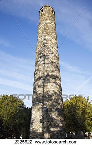 Stock Image - Low angle view of a round tower against a blue sky and a cemetery; Glendalough county wicklow ireland. Fotosearch
