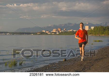Stock Image - Man jogging on the beach next to the tony knowles coastal trail during high tide at sunset; Anchorage alaska united states of america. Fotosearch