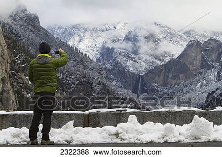 Stock Photo - Man wearing a green down jacket as seen from the back taking a picture of yosemite valley and bridal veil falls; Yosemite california united states of america. Fotosearch
