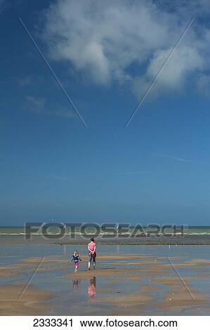 Stock Image - Mother and young daughter walking across sandy beach at low tide; Granville brittany france. Fotosearch