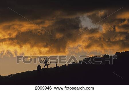 Stock Photograph - Mountain Biker Carries Bike Up Rocky Slope, Sunset Clouds Behind, Whistler, Bc Canada. Fotosearch