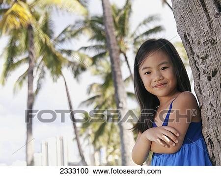 Stock Image - Portrait of a young girl leaning against the trunk of a tree; Honolulu oahu hawaii united states of america. Fotosearch