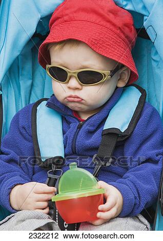 Stock Image - Portrait of young boy wearing a red hat and sunglasses sitting in a stroller with a grumpy look on his face; Anchorage alaska united states of america. Fotosearch