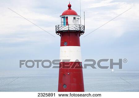 Stock Image - Red and white lighthouse along the coast with a sailboat in the distance near westkapelle; Zealand netherlands. Fotosearch