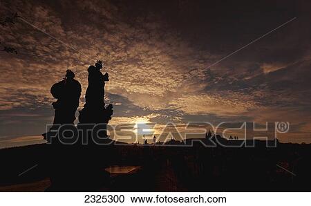 Stock Image - Silhouette of two statues and the skyline in the distance at sunset; Prague czech republic. Fotosearch