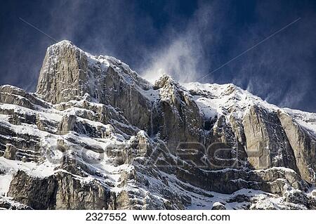 Stock Image - Snow covered mountain peak with blowing snow off the peak and blue sky; Banff alberta canada. Fotosearch