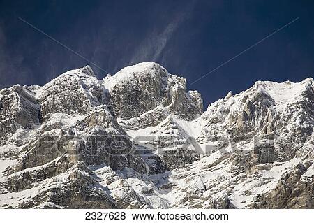 Stock Photo - Snow covered mountain peak with snow blowing off peak and blue sky; Banff alberta canada. Fotosearch