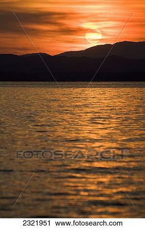 Sun Setting Over A Silhouetted Mountain Reflecting On A Lake; Bardolino Veneto Italy View Large Photo Image Stock Image - Sun Setting Over A Silhouetted Mountain Reflecting On A Lake; Bardolino Veneto Italy. Fotosearch