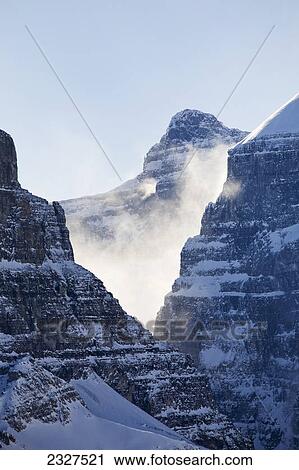 Stock Image - Sunlit snow blowing off a mountain peak in a v shaped rock cliff with blue sky; Lake louise alberta canada. Fotosearch