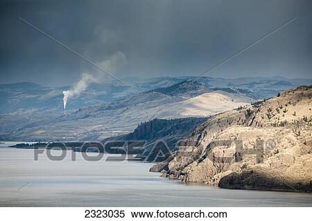 Stock Photography - The dry hills of kamloops with a smokestack spewing pollution in the distance; Kamloops british columbia canada. Fotosearch
