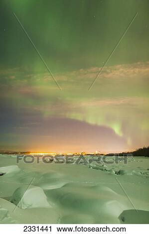 Stock Image - The northern lights shine above the anchorage city skyline and light the snow green in this nighttime view from the tony knowles coastal trail in winter; Anchorage alaska united states of america. Fotosearch