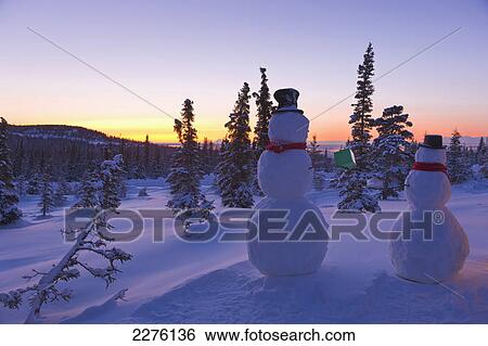 Stock Photograph - Two snowmen exchanging gifts in front of a snow covered spruce forest after sunset with chugach mountains; Anchorage alaska united states of america. Fotosearch