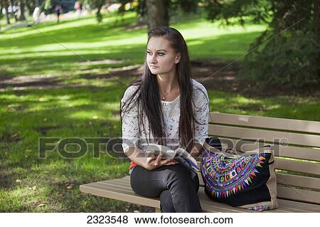 Stock Photo - Young woman reading in a park sitting on a park bench; Edmonton alberta canada. Fotosearch