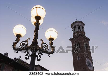 Stock Image - A clock tower and illuminated lamp post in Piazza Delle Erbe at dusk; Verona, Italy. Fotosearch