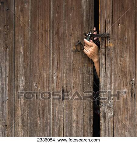 Picture - A hand reaching out a wooden door to unlock a padlock. Fotosearch