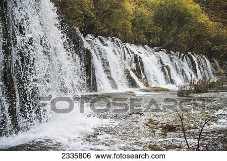 Stock Photograph - A long line of waterfalls along a cliff with trees growing on the top edge of the cliff. Fotosearch