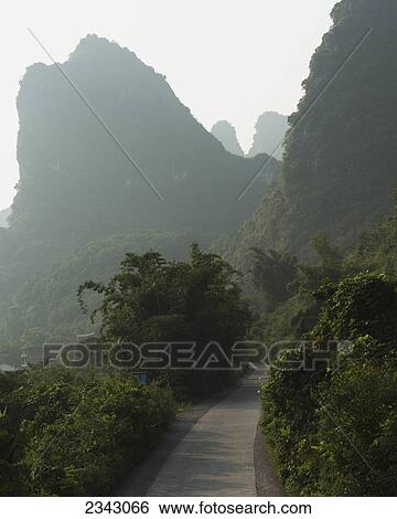 Stock Photograph - A path going through a landscaped area with mountain peaks and fog; Yangshuo, Guangxi, China. Fotosearch