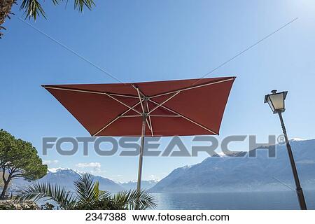 A patio umbrella and lamp post against a blue sky with a view of Lake Maggiore and the Swiss Alps; Brissago, Ticino, Switzerland View Large Photo Image Stock Photo - A patio umbrella and lamp post against a blue sky with a view of Lake Maggiore and the Swiss Alps; Brissago, Ticino, Switzerland. Fotosearch