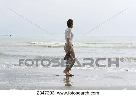 Stock Image - A woman walking on the wet beach carrying her shoes and looking out at the ocean; Rimini, Emilia-Romagna, Italy. Fotosearch