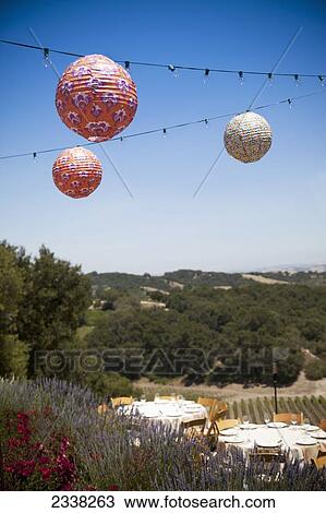 Stock Image - Paper lanterns hanging from strings of lights over tables set for dinner; California, United States of America. Fotosearch