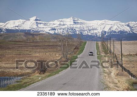 Stock Photo - Paved road with single vehicle along fields with snow covered mountians and blue sky in the background; Longview, Alberta, Canada. Fotosearch